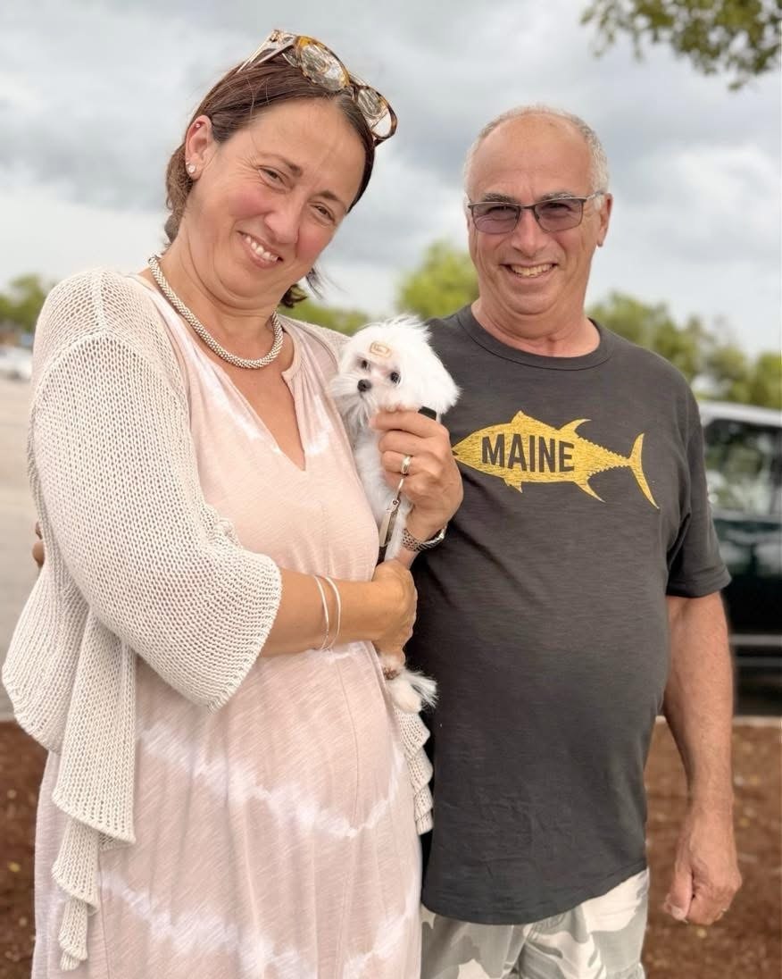 Couple standing outdoors on an overcast day holding a small white Maltese puppy between them