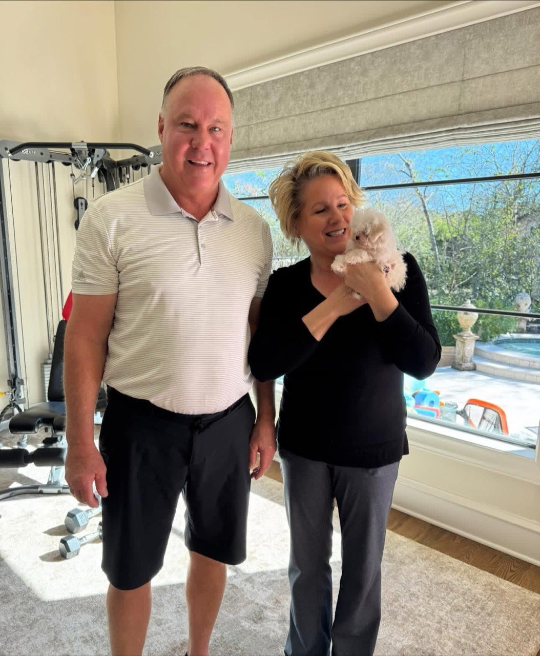 Two people standing beside a bright window at home while one of them holds a tiny white Maltese puppy