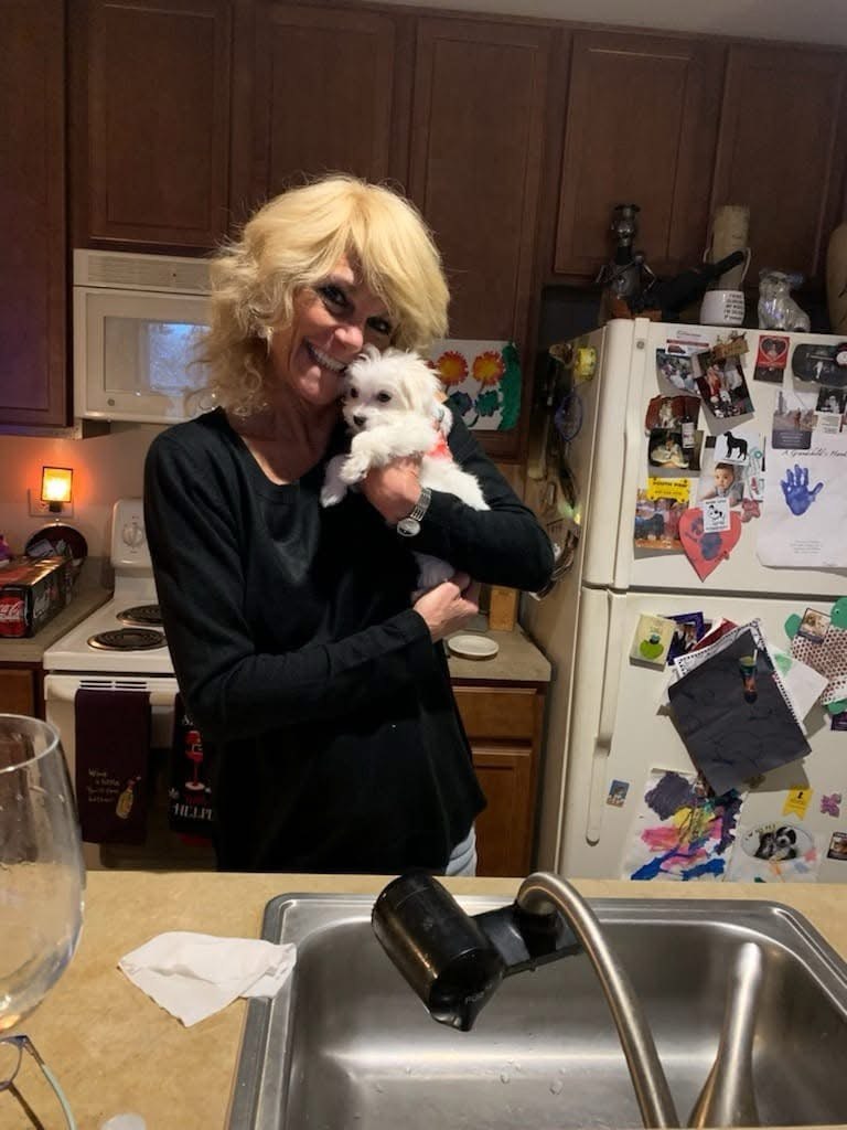 Smiling woman cuddling a tiny white Maltese puppy against her cheek in a home kitchen